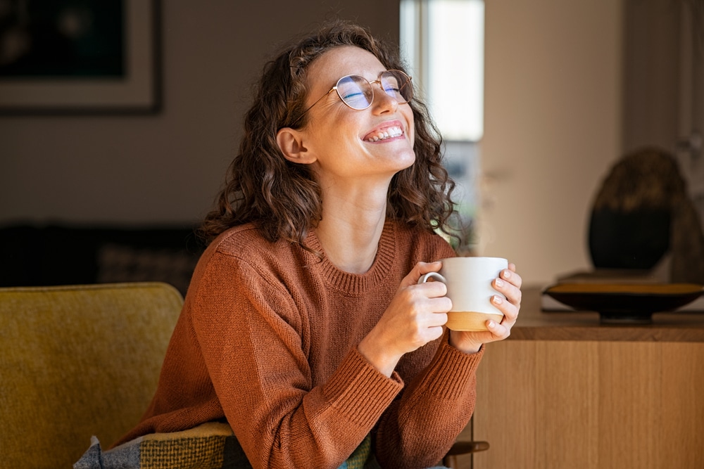 A woman leaning forward on a couch smiling and holding a cup of coffee in her hands.