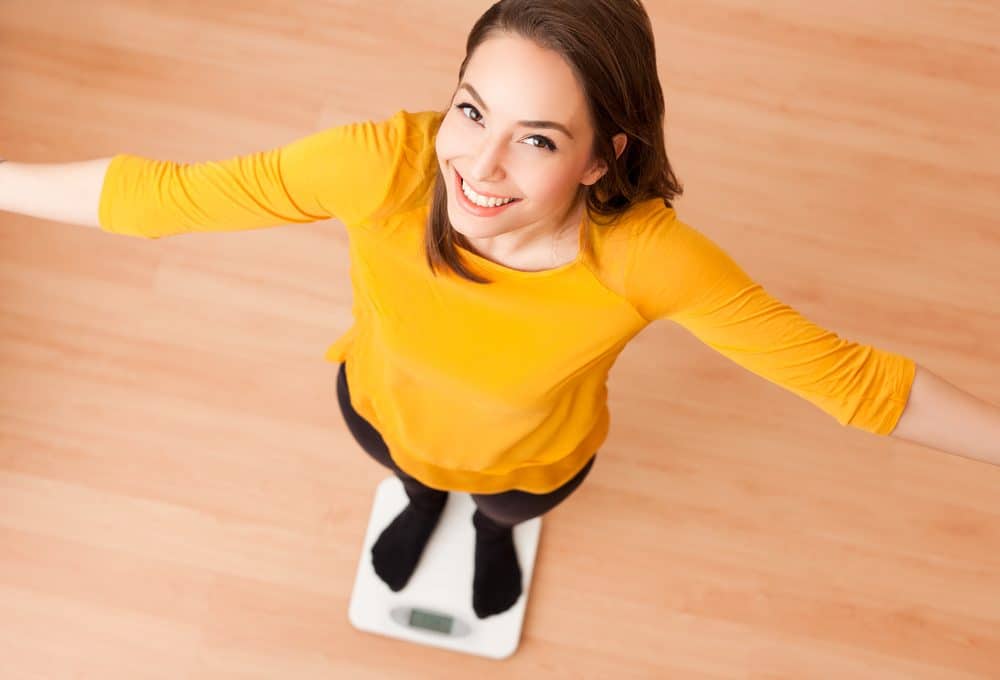 A woman standing on a scale with her arms out and smiling up at the camera.