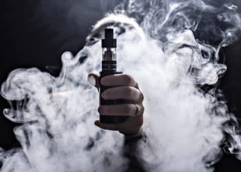 Someone holding out a vape toward the camera with smoke all around their hand and arm.