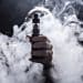 Someone holding out a vape toward the camera with smoke all around their hand and arm.