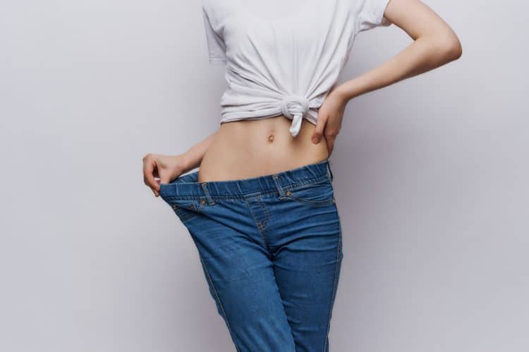 A woman only shown from her torso down to her legs, pulling her jeans out to show how much skinnier she is than them.